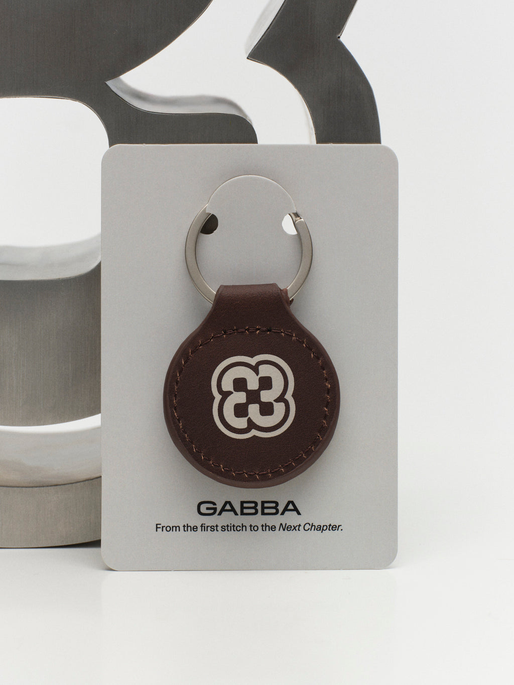 The Gabba Leather Key Ring - Brown features a white Gabba Brand Mark on brown leather, attached to a polished metal ring and displayed on a card reading “GABBA From the first stitch to the Next Chapter.”.